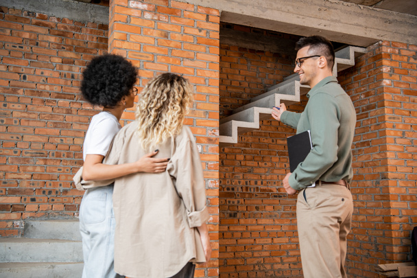 agent immobilier pointant vers les escaliers et montrant la maison avec intérieur inachevé pour lesbienne couple interracial - Photo, image
