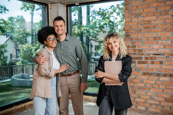 successful realtor with folder and cheerful multiethnic couple looking at camera in new cottage - Photo, Image