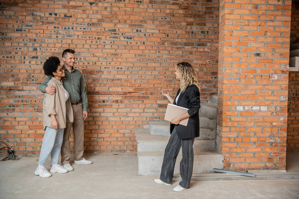 real estate broker with folder showing unfinished interior of new house to interracial couple - Photo, Image