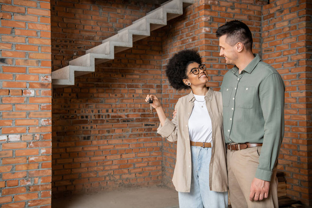 überglückliche Afroamerikanerin hält Schlüssel in der Hand und schaut Mann in neuem Eigenheim an - Foto, Bild