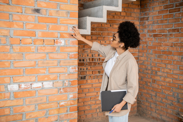 Afrikaans Amerikaans makelaar met map aanraken baksteen muur in huis met onafgewerkte interieur - Foto, afbeelding