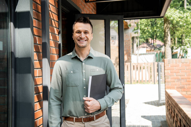 successful and happy real estate agent with folder looking at camera near modern cottage - Photo, Image