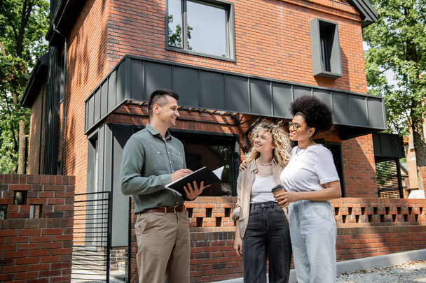 positive real estate agent talking to interracial lesbian couple with coffee to go near city cottage - Photo, Image