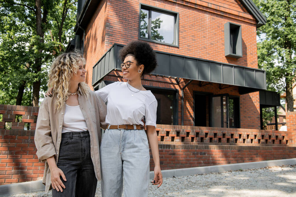 joyful multiethnic lesbian couple looking at each other next to private modern cottage - Foto, imagen