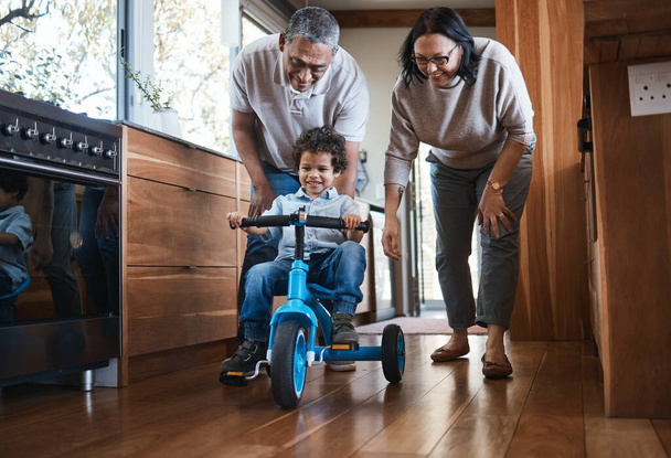 Grandparents, teaching child to ride and bike in kitchen, home or childhood memory of learning with elderly people. Kid, grandmother and grandfather with bicycle and cycling on training wheels. - Фото, изображение