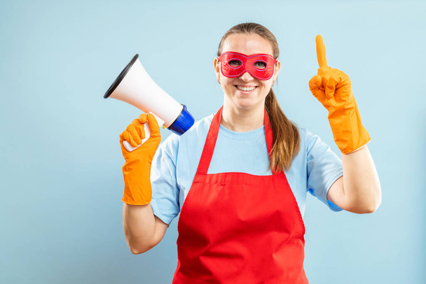 Schoonmaakster in rood heldenmasker, handschoenen en schort met megafoon op blauwe achtergrond. Informatie over schoonmaakdiensten - Foto, afbeelding