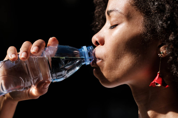Detailní záběr černošky, jak pije vodu z láhve. Zdravý životní styl a koncepce nealkoholických nápojů. - Fotografie, Obrázek