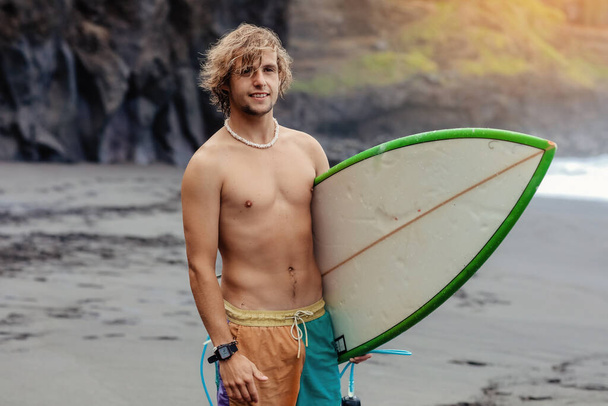 Komea istuvuus nuori vaalea mies, jolla on mallina surffilauta odottaa aallon surffata paikalla meren rannalla mustalla hiekalla ja katsoo kameraa. Käsite urheilu, kunto, vapaus, onnellisuus, uusi moderni elämä. - Valokuva, kuva