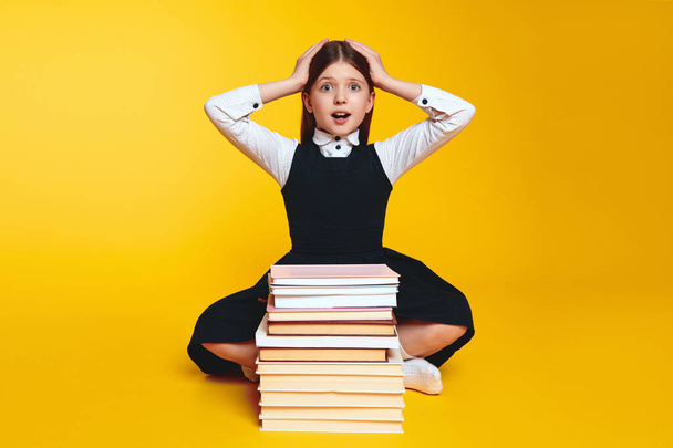 Astonished girl in school uniform keeping hands near head while sitting near stack of books, isolated over yellow background. Back to school concept - Foto, afbeelding