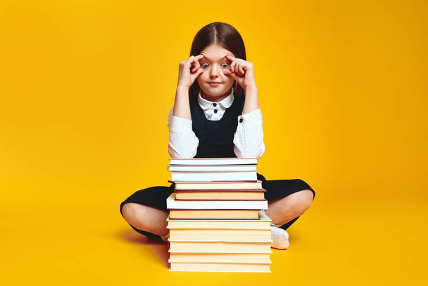 Σοκαρισμένη μαθήτρια ανοίγει τα μάτια της με δάχτυλα και κοιτάζει ένα σωρό βιβλία, που δεν είναι έτοιμα για μελέτη, κάθεται απομονωμένη σε κίτρινο φόντο - Φωτογραφία, εικόνα