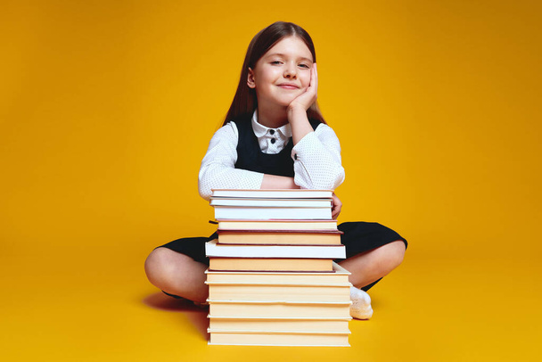 Young girl wearing uniform holding hands on stack of books while looking at camera, sitting isolated over yellow background - Photo, image