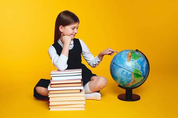 Jolie écolière tittle pointant vers le monde globe tandis que assis près de tas de livres, isolé sur fond jaune, espace de copie. Concept de retour à l'école - Photo, image
