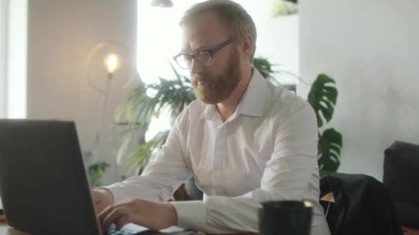 Γενειοφόρος άνδρας με γυαλιά και λευκό επίσημο πουκάμισο που εργάζονται σε φορητό υπολογιστή στο γραφείο σε άνετο γραφείο - Πλάνα, βίντεο