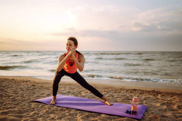 Beautiful young woman in sportswear practicing stretching on yoga mat alone on wild beach. Healthy lifestyle concept - Valokuva, kuva