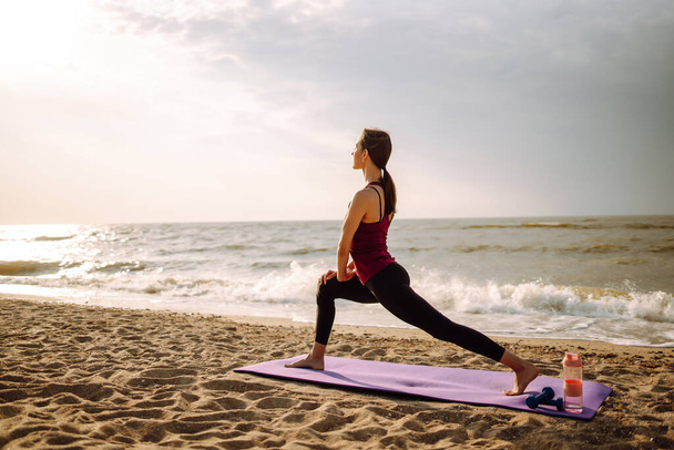 Beautiful young woman in sportswear practicing stretching on yoga mat alone on wild beach. Healthy lifestyle concept - Zdjęcie, obraz