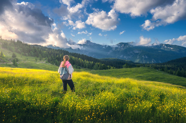 Girl on the hill with yellow flowers and green grass in beautiful alpine mountain valley at sunset in summer. Landscape with young woman in alps, trees, sky with clouds. Travel and Hiking. Slovenia - Foto, imagen