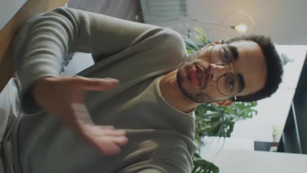 Fotografía vertical del hombre birracial gen Z en gafas y atuendo casual hablando en cámara mientras filmaba vlog en casa o web llamando - Metraje, vídeo