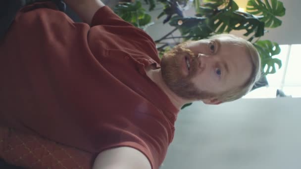 Függőleges lövés vörös hajú szakállas férfi gazdaság smartphone kinyújtott kar és beszél a kamera keresztül videohívás otthon - Felvétel, videó
