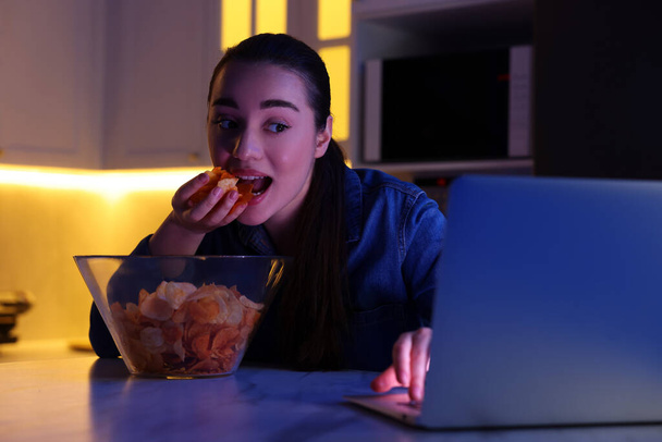 Young woman eating chips while using laptop in kitchen at night. Bad habit - Φωτογραφία, εικόνα