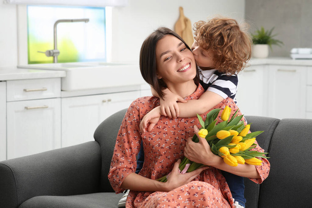 Little son kissing and congratulating his mom with Mother`s day at home. Woman holding bouquet of yellow tulips - Zdjęcie, obraz