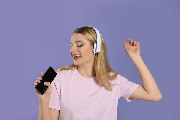 Happy woman in headphones enjoying music and singing into smartphone on violet background - Фото, изображение