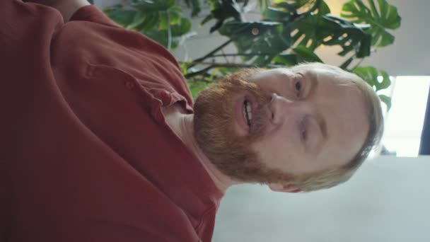 Κάθετη λήψη του κοκκινομάλλα γενειοφόρος άνδρας χαιρετώντας και μιλώντας στην κάμερα, ενώ κουβεντιάζοντας μέσω βιντεοκλήσης στο σπίτι - Πλάνα, βίντεο