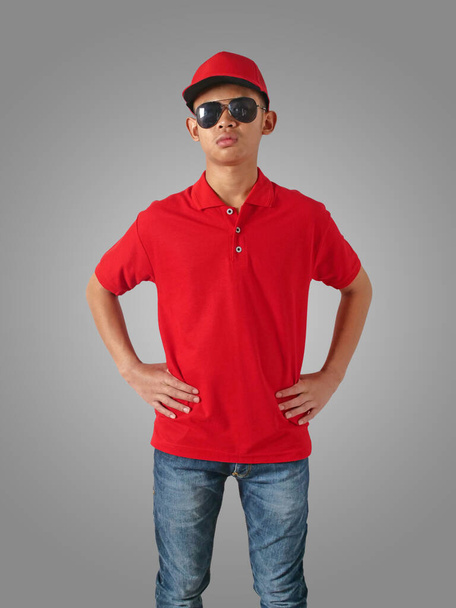 Young Asian teenage boy wearing red collared shirt, hat and sunglasses standing with hands on hip over grey background, t-shirt template mock up - Foto, Imagen