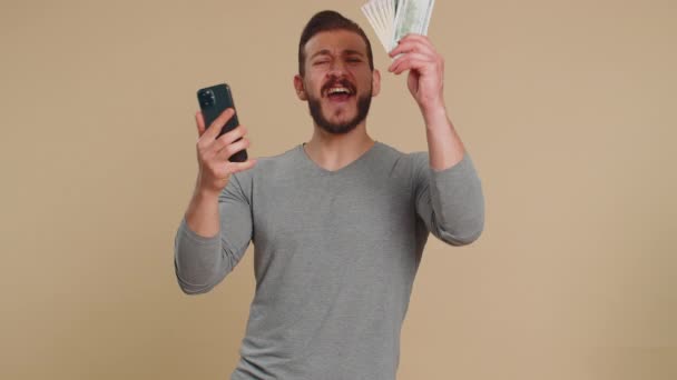 Lebanese man looking smartphone display sincerely rejoicing win, receiving money dollar cash banknotes, success lottery jackpot luck, income salary wage. Middle eastern guy on studio beige background - Footage, Video