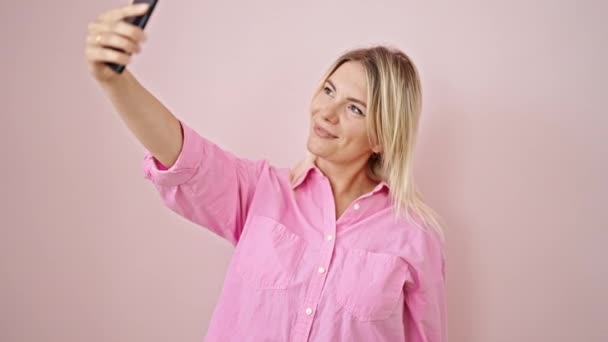 jonge blonde vrouw glimlachen zelfverzekerd maken selfie door de smartphone over geïsoleerde roze achtergrond - Video