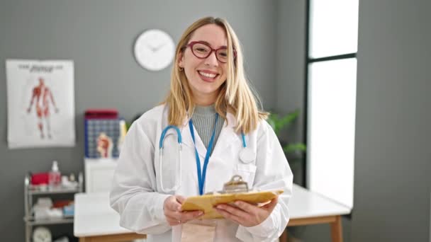 Νεαρή ξανθιά γυναίκα γιατρός χαμογελά αυτοπεποίθηση κρατώντας πρόχειρο στην κλινική - Πλάνα, βίντεο