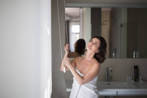 Middle-aged woman opening white roller blinds on window in a modern apartement - Foto, afbeelding