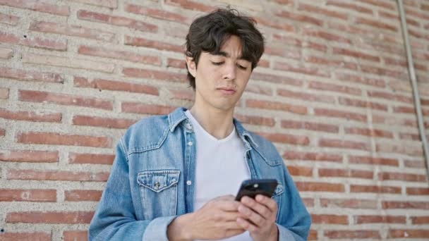 Fiatal spanyol férfi használ okostelefon komoly kifejezés elszigetelt tégla háttér - Felvétel, videó