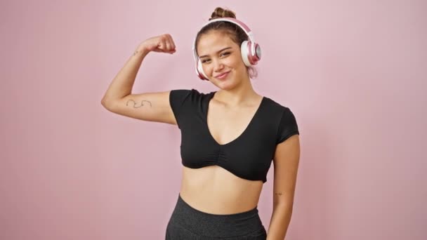 Giovane bella donna ispanica ascoltare musica facendo gesto forte con le braccia su sfondo rosa isolato - Filmati, video