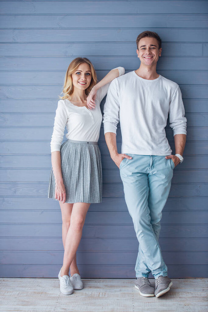 Full length portrait of beautiful young couple in casual clothes looking at camera and smiling, standing against gray wall - Zdjęcie, obraz