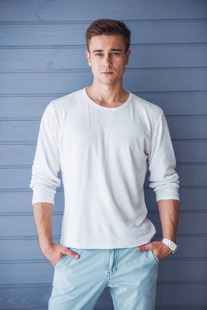 Portrait of handsome young man in casual clothes looking at camera, standing with hands in pockets against gray wall - Foto, afbeelding
