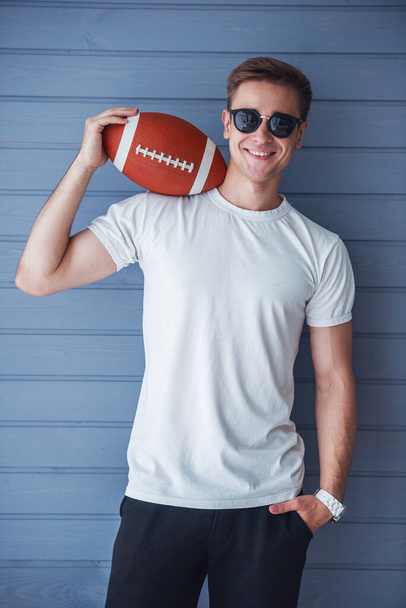 Przystojny młody człowiek w casual ubrania i okulary przeciwsłoneczne trzyma amerykański piłkę nożną, patrząc na aparat i uśmiechnięty, stoi na szarej ścianie - Zdjęcie, obraz