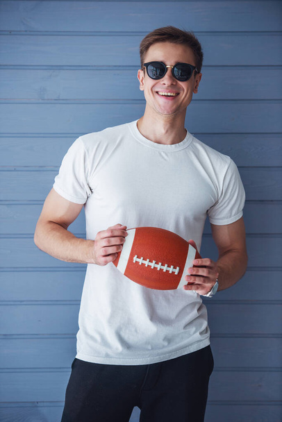 Handsome young man in casual clothes and sun glasses is holding an American football ball, looking at camera and smiling, standing against gray wall - Fotografie, Obrázek