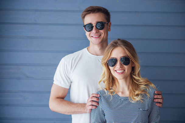 Portrét krásného mladého páru v příležitostných šatech a slunečních brýlích, které se dívají na kameru a usmívají se, stojící proti šedé zdi - Fotografie, Obrázek