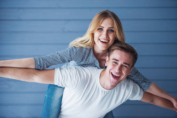 Portrait of beautiful young couple in casual clothes looking at camera and smiling, standing against gray wall. Girl is sitting pickaback - Photo, Image