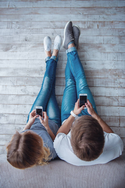 Vista superior de la hermosa pareja joven en ropa casual usando teléfonos inteligentes mientras está sentado en el suelo contra la pared gris
 - Foto, Imagen