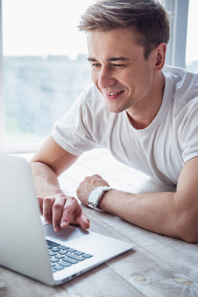 Pohledný mladý obchodník v příležitostných šatech používá laptop a usmívá se, zatímco leží doma na podlaze před okny - Fotografie, Obrázek