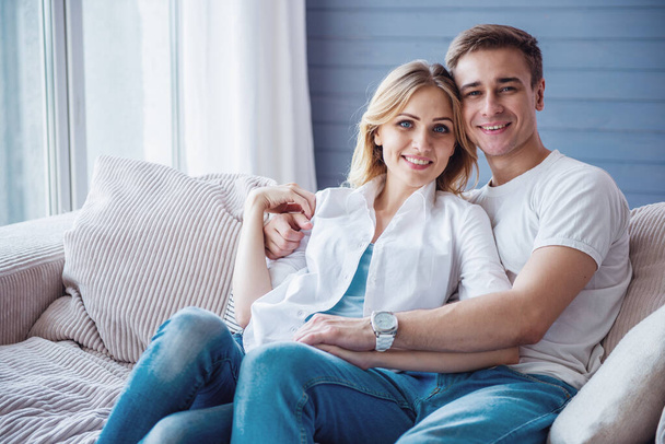 Krásný mladý pár v příležitostných šatech se drží za ruce, dívá se na kameru a usmívá se, zatímco sedí na gauči doma - Fotografie, Obrázek