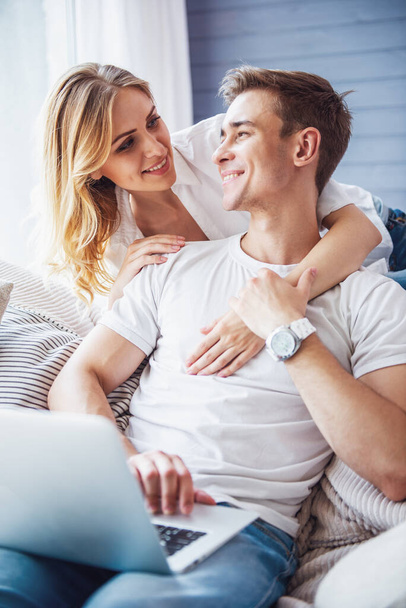 Handsome young businessman in casual clothes is using a laptop, sitting on couch at home while his girl is hugging him behind. Both are looking at each other and smiling - 写真・画像