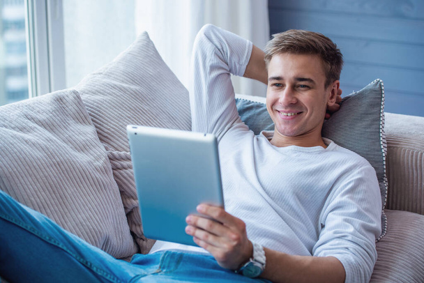 Komea nuori liikemies rennoissa vaatteissa käyttää digitaalista tablettia ja hymyilee kotona sohvalla.
 - Valokuva, kuva