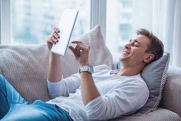 Guapo joven hombre de negocios en ropa casual está utilizando una tableta digital y sonriendo mientras está acostado en el sofá en casa
 - Foto, Imagen
