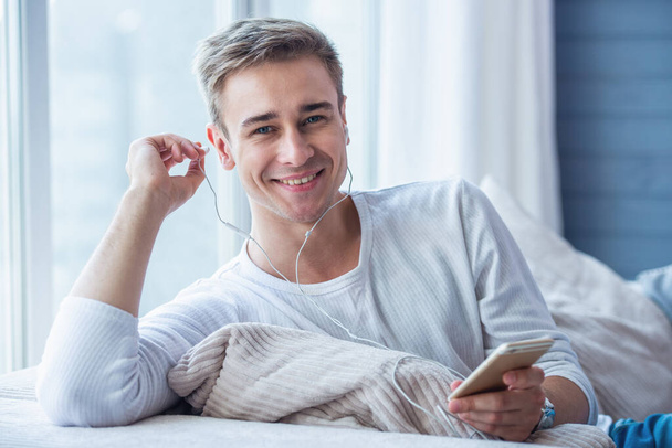 Guapo joven hombre de negocios en los auriculares está escuchando música usando un teléfono inteligente, mirando a la cámara y sonriendo mientras está sentado en el sofá en casa
 - Foto, Imagen
