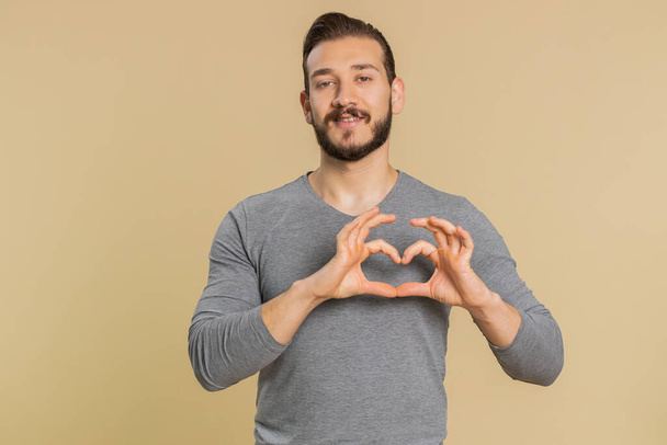 I love you. Smiling happy lebanese young man makes heart gesture demonstrates love sign expresses good romantic feelings and sympathy symbol. Middle eastern guy isolated on beige studio background - Photo, Image