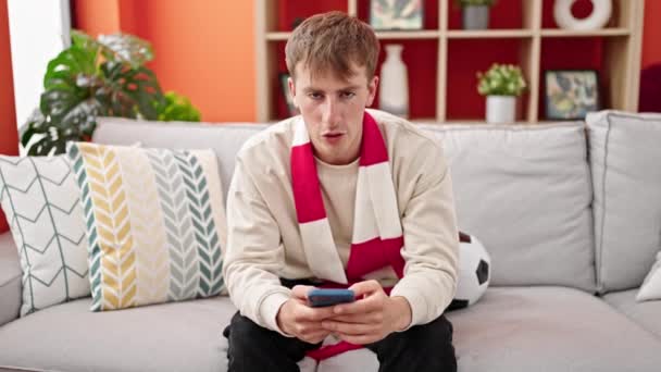 Joven hombre caucásico apoyando equipo de fútbol usando teléfono inteligente en casa - Metraje, vídeo