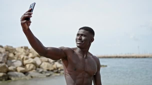 Afrikanischer Mann steht hemdlos und macht Selfie mit Smartphone am Strand - Filmmaterial, Video