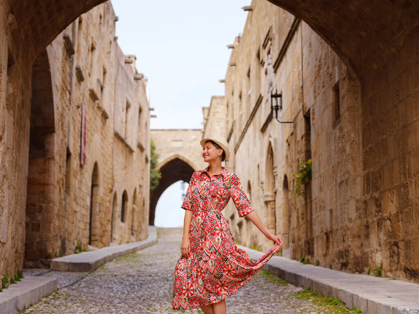 summer trip to Rhodes island, Greece. Young Asian woman in ethnic red dress walks Street of Knights of Fortifications castle. female traveler visiting southern Europe. Unesco world heritage site. - Photo, Image